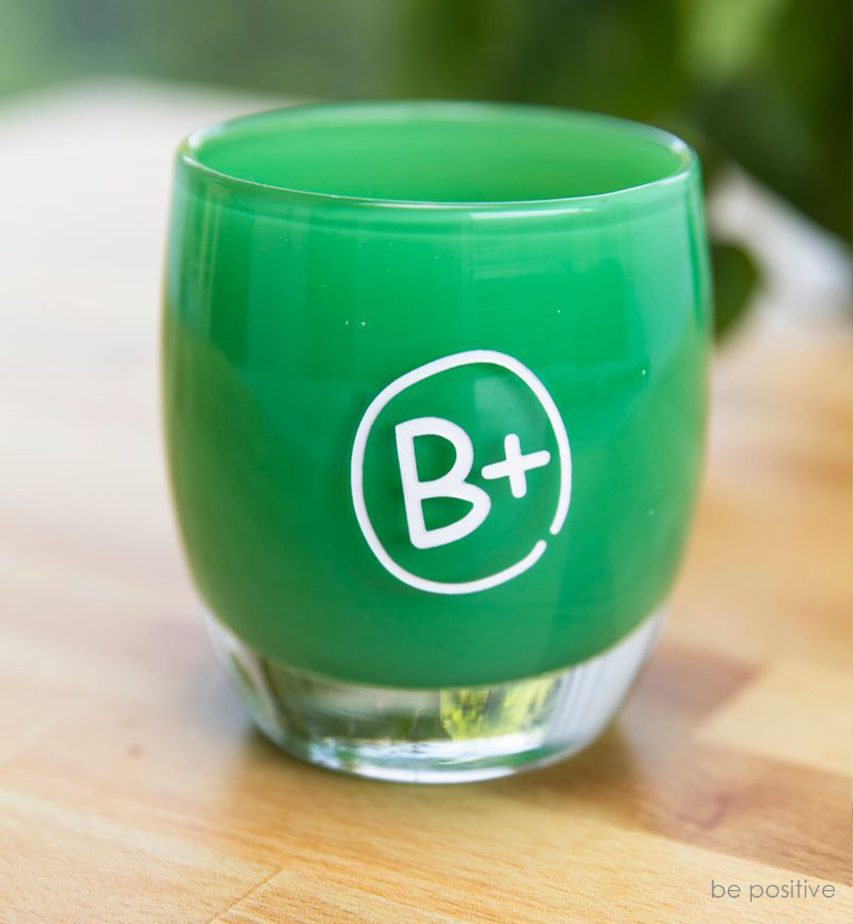 emerald green hand-blown glass votive candle holder with The B+ Foundation logo hand-etched in white on the side. sitting on a light wood table.