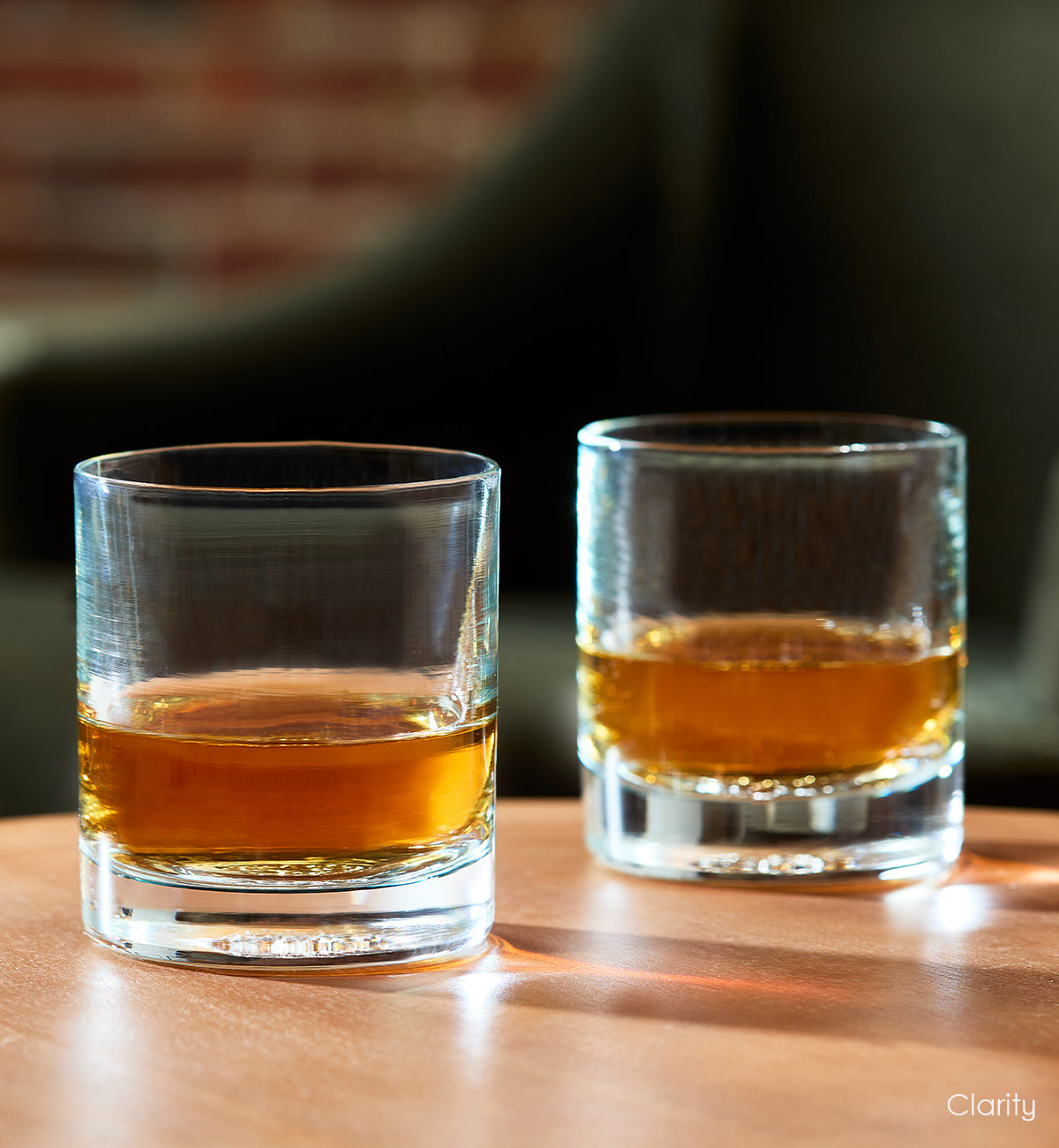 two Clarity rockers, clear with light blue hues hand-blown glass lowball drinking glasses with dark liquid inside, sitting on light wood table with green chair in background.