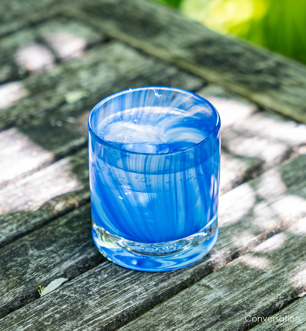 conversation, raked blue hand-blown glass lowball drinking glass on a wood patio table sitting in sunlight and shadows.
