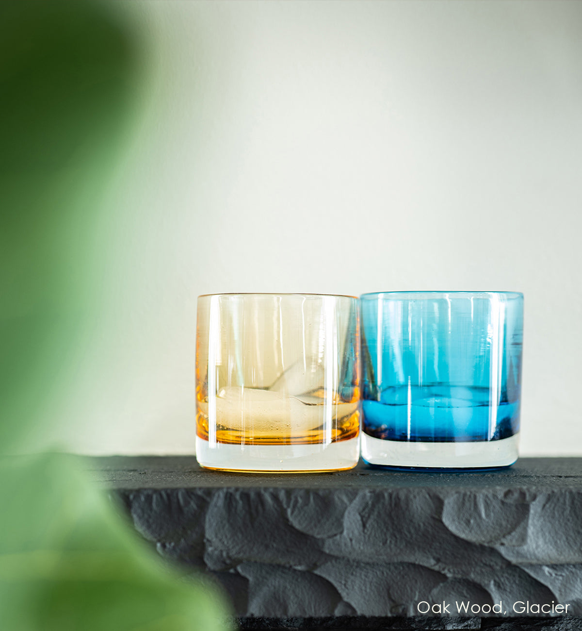Oak Wood and Glacier rockers, light blue transparent hand-blown glass lowball drinking glass next to light brown hand-blown drinking glass on top of a charcoal table with white background.