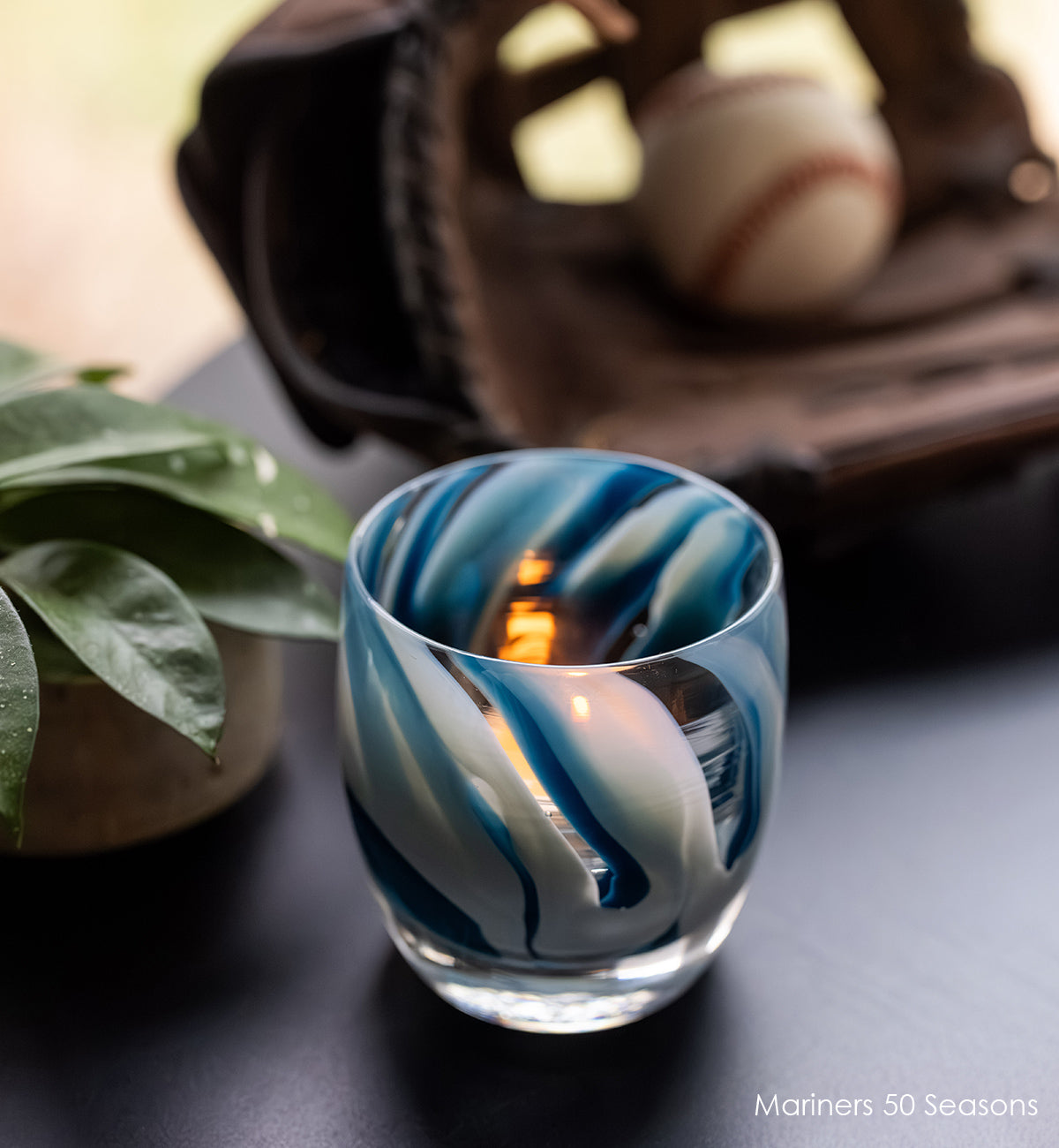 Mariners 50 Seasons glass votive candle holder with blue and white swirl design on a dark surface with baseball and glove in background.