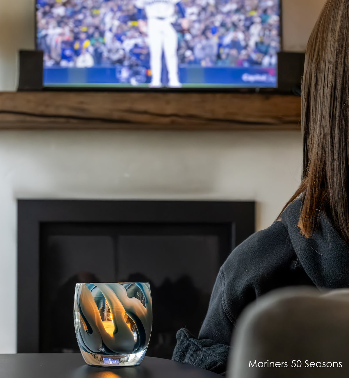 Person watching a sports game on TV with a Mariners 50 Seasons glass votive candle holder with blue and white swirl design on a dark surface with baseball and glove in background.