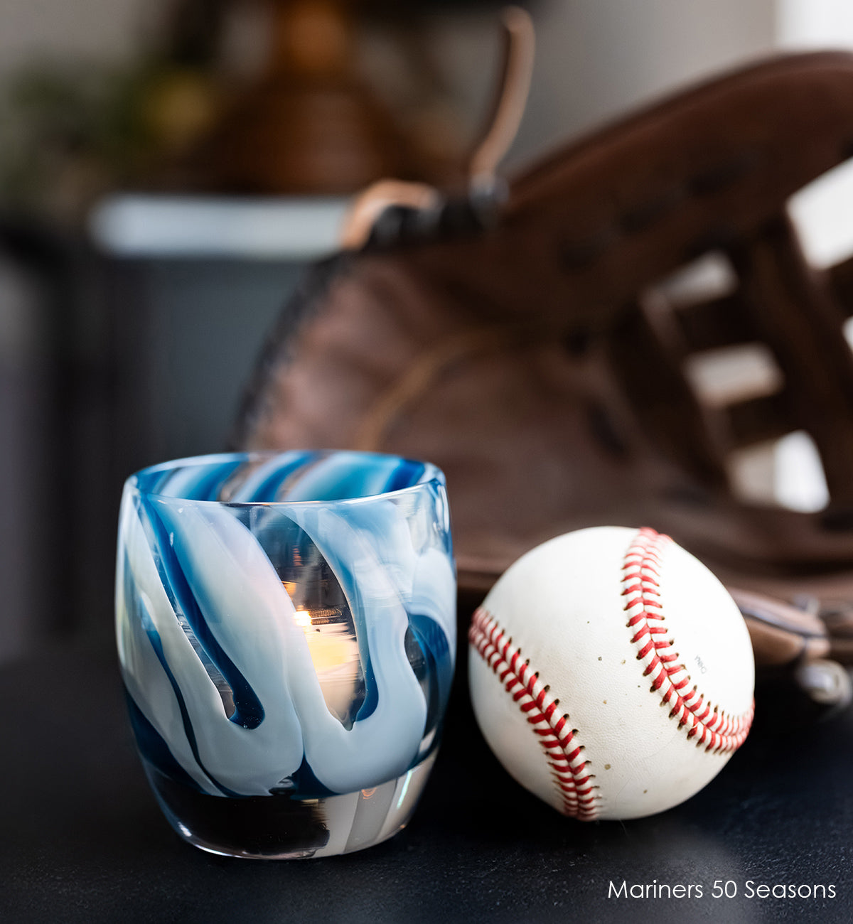 Mariners 50 Seasons glass votive candle holder with blue and white swirl design on a dark surface with baseball and glove in background.