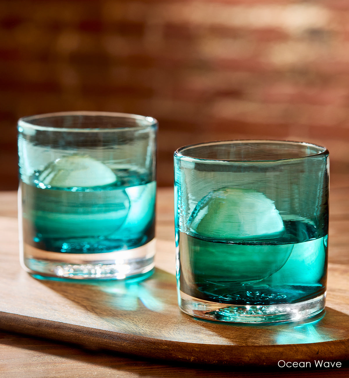 two Ocean Wave rockers, teal blue transparent hand-blown lowball drinking glasses with liquid and ice on a dark wood table with brick wall background.