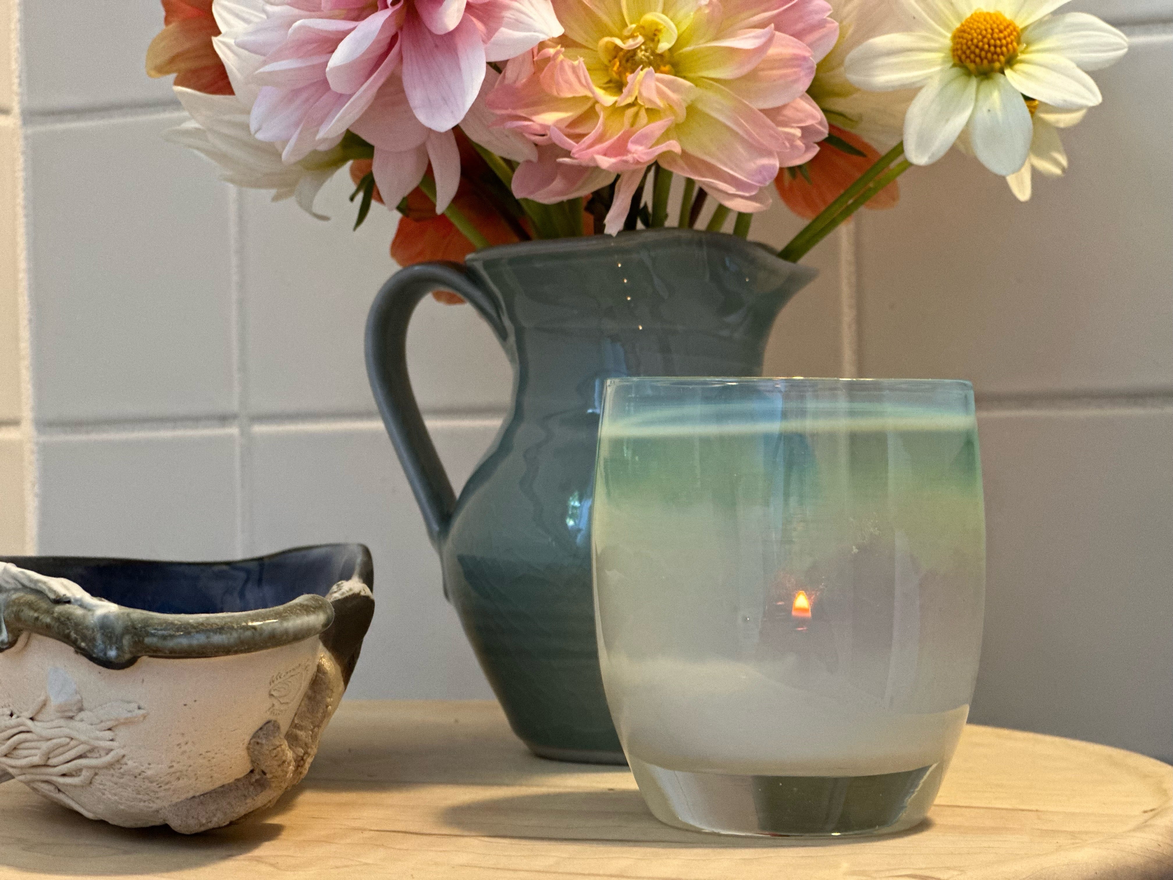 enchanted, misty green with metallic luster hand-blown glass votive candle holder on a wood side table with teal flower vase and beachy bowl.
