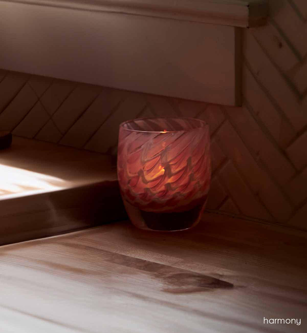 harmony, pink feathered texture hand-blown glass votive candle holder. Glowing a flame on a wooden counter.