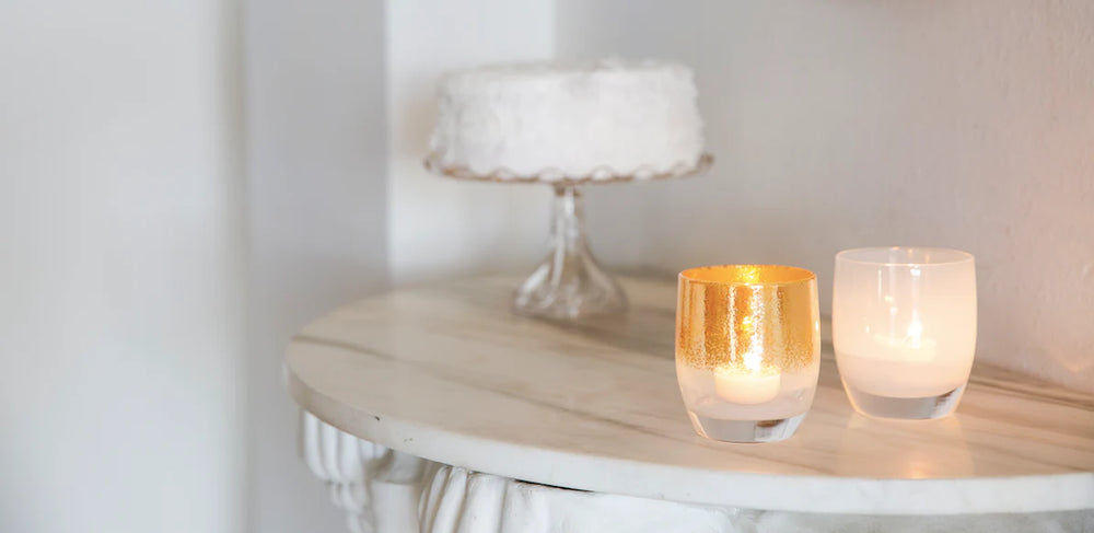 grace gold on white, and celebrate white, hand-blown glass votive candle holders with lit tealights inside on a marble side table with cake.