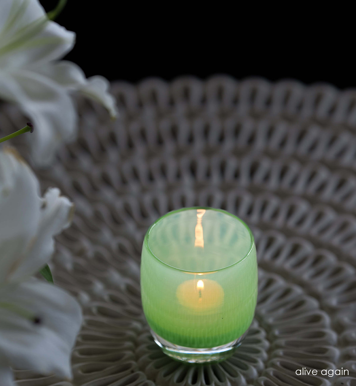 alive again vibrant green, hand-blown glass votive candle holder on a wicker table next to lilies.