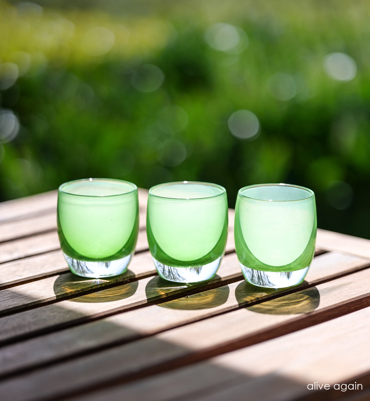 three alive again vibrant green, hand-blown glass votive candle holders on patio table sitting in the sunshine.