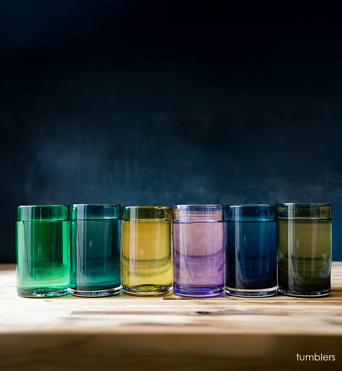 all tumblers hand-blown drinking glasses filled with water. on wood table navy background.