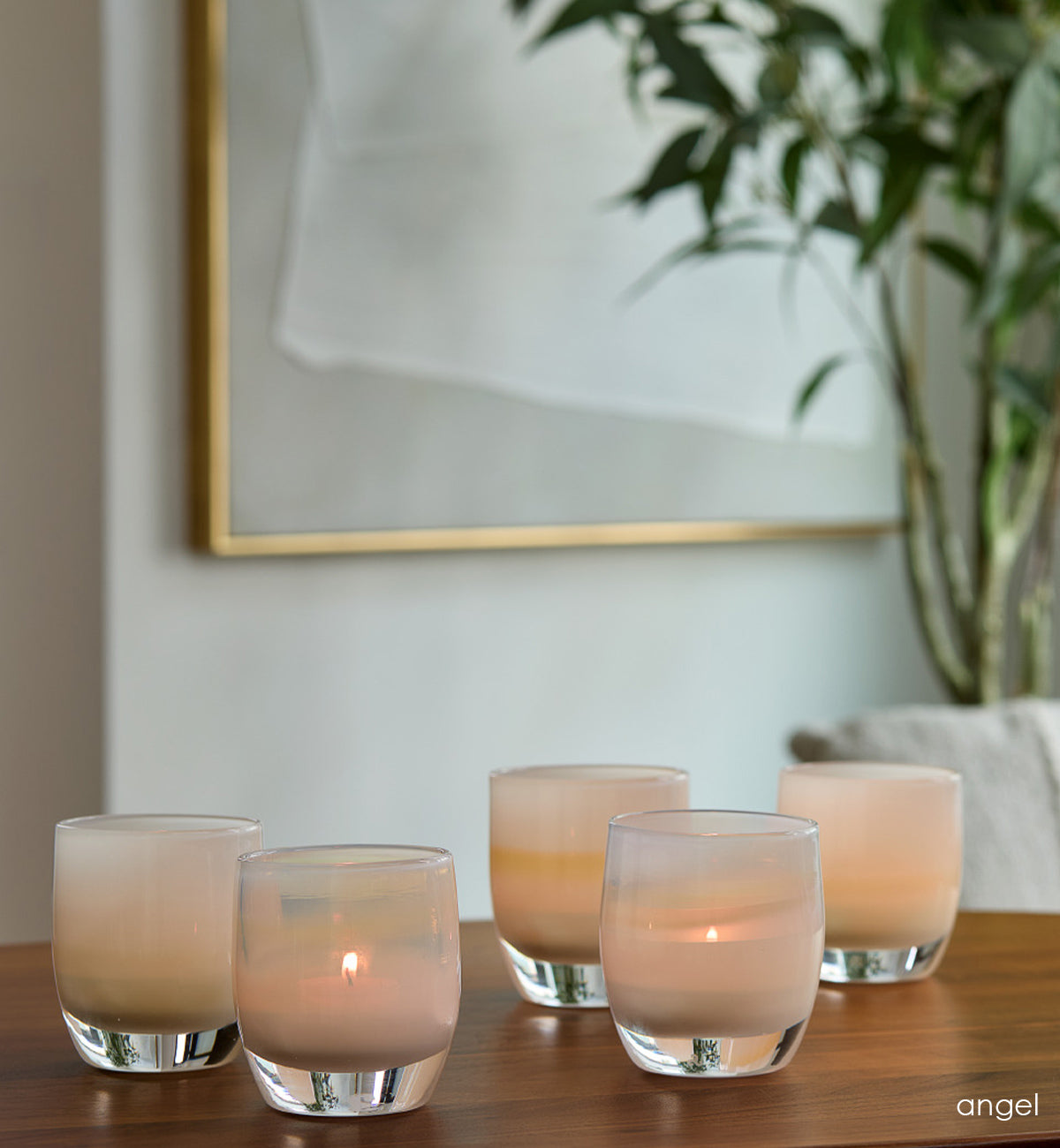 grouping of five angel light yellow, hand-blown glass votive candle holders on a wood table with a plant, framed art and chair in background.