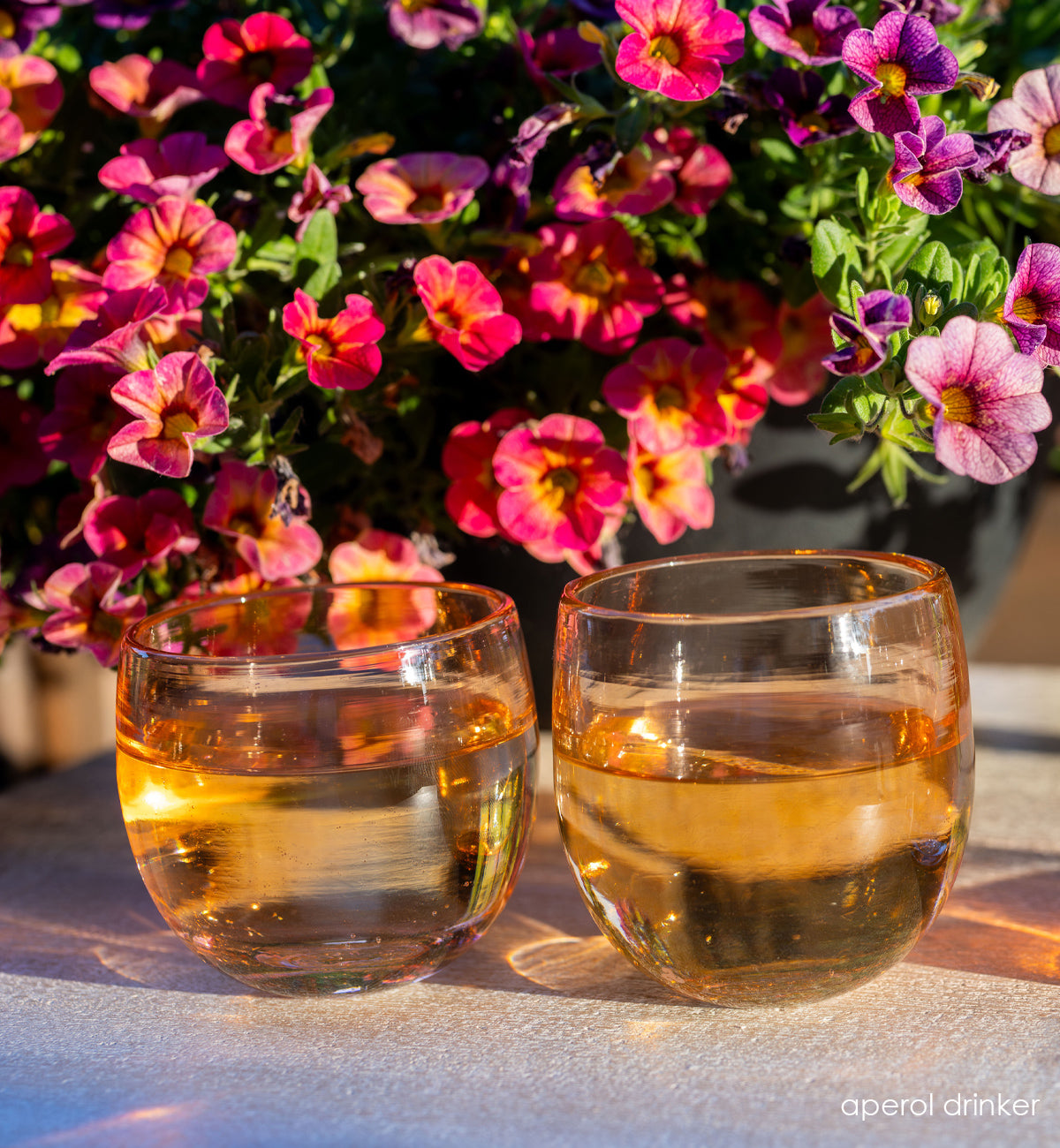 aperol set, two coral hand-blown drinking glasses filled with water. pink and purple flowers in the background.