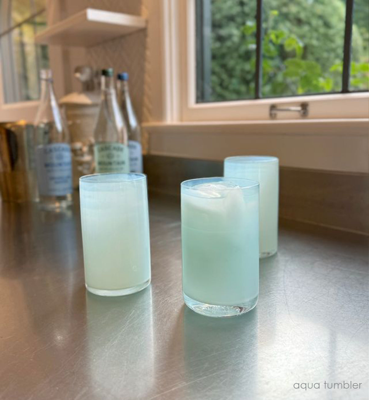 three aqua tumblers, translucent seafoam green hand-blown water glasses with liquid and ice on a kitchen counter with sparkling water bottles and a window in background.