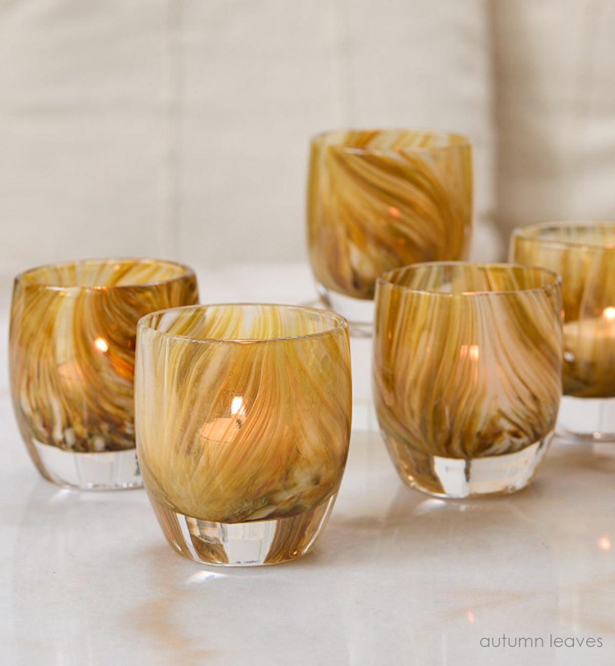 several autumn leaves, yellow brown raked hand-blown glass votive candle holders with lit tealights on a cream marble surface.