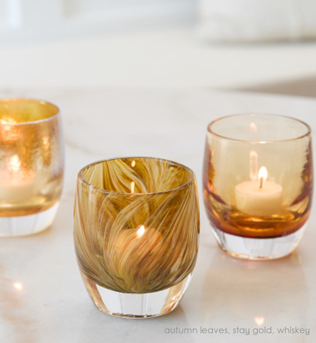autumn leaves, yellow brown raked hand-blown glass votive candle holder with stay gold and whiskey on a marble surface.