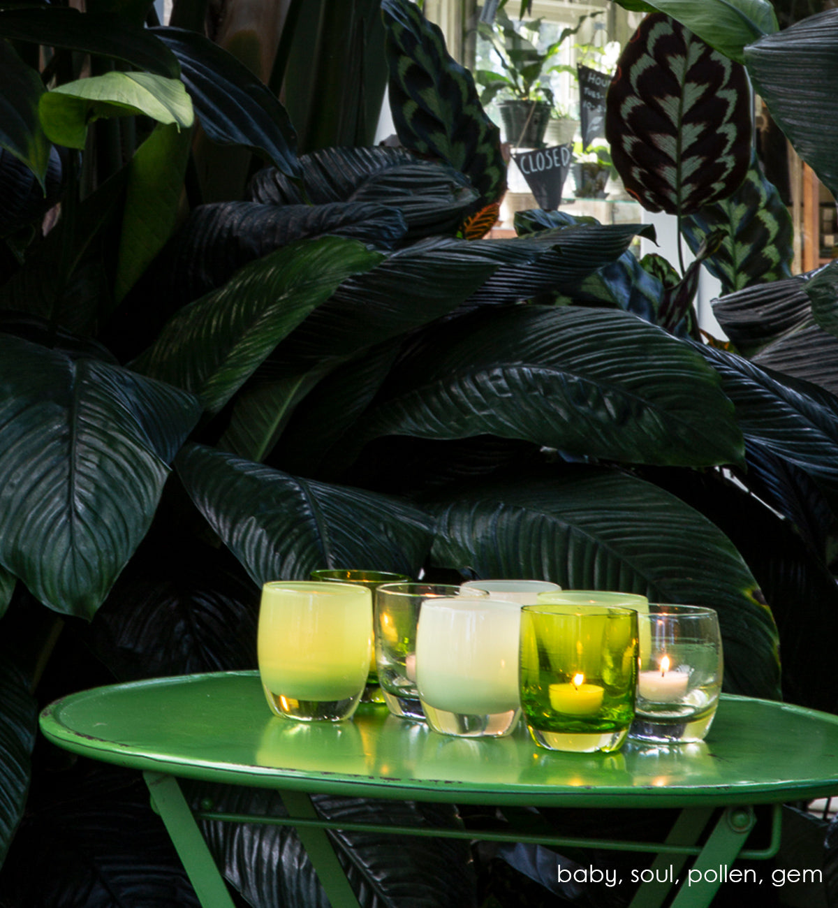 baby light green opaque, soul iridescent translucent, bud pale green opaque, gem green transparent, hand-blown glass votive candle holder iwth soul pollen, and gem on a green metal patio table.