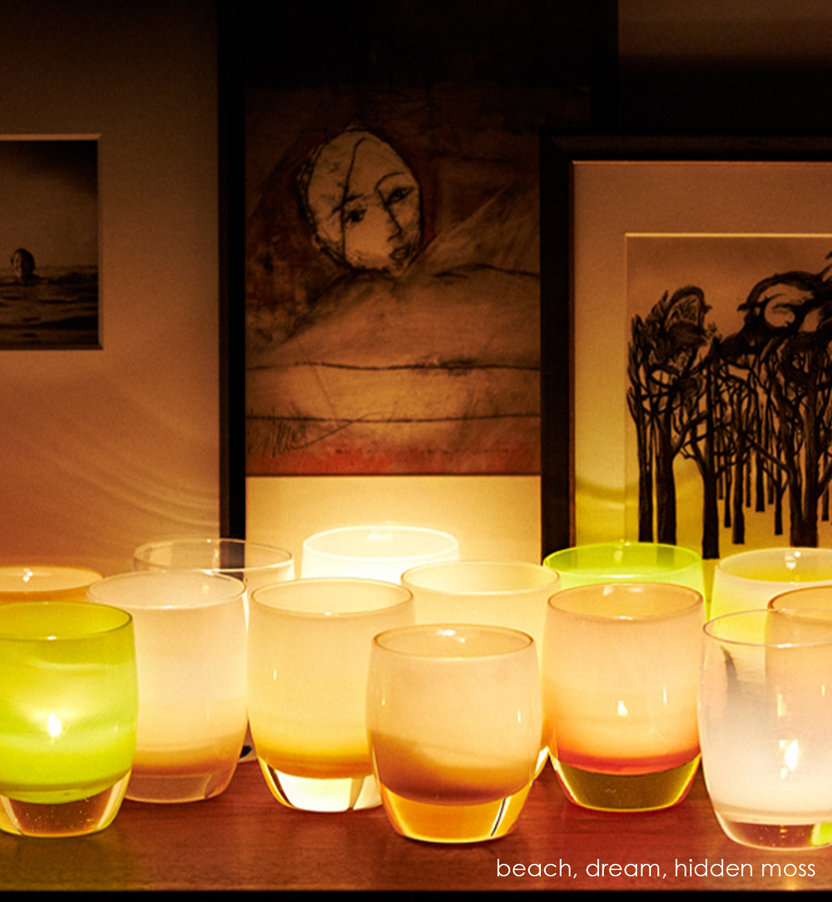 hidden moss, pear green hand-blown glass votive candle holders on a wood table with artwork behind lit with several beach and dream.