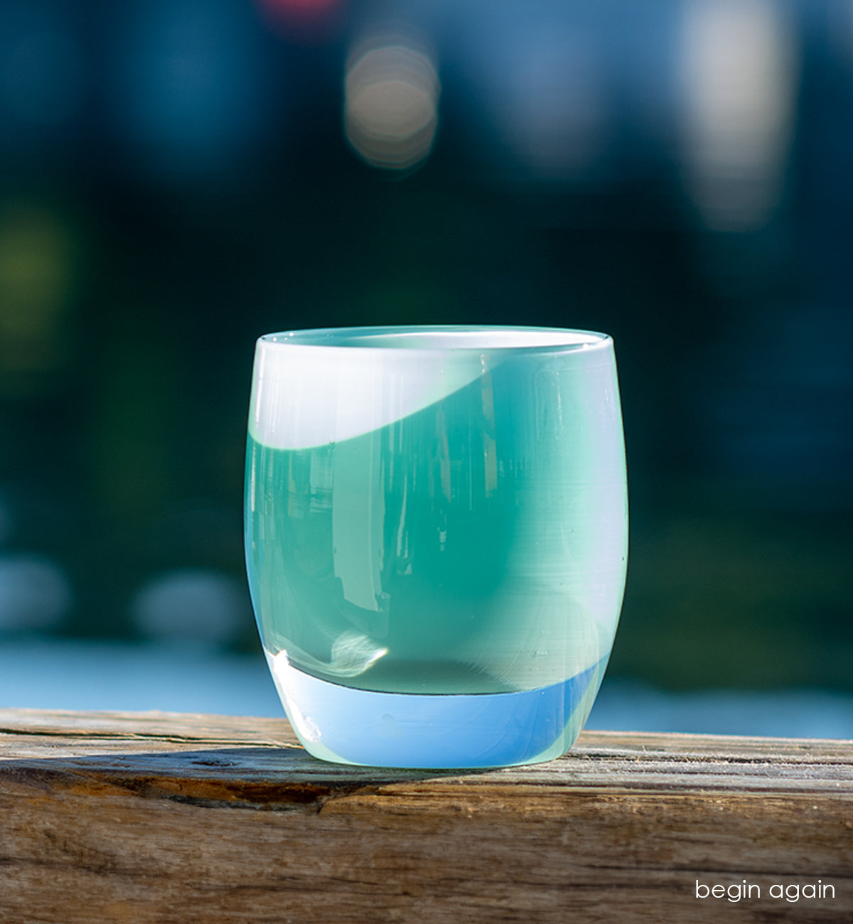 begin again light teal, hand-blown glass votive candle holder sitting in sunlight on wood perch with a body of water and boat blurred in background.