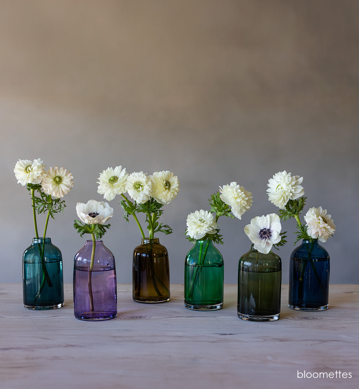 transparent blue hand-blown glass flower bud vase with white flowers on wood table and gray background