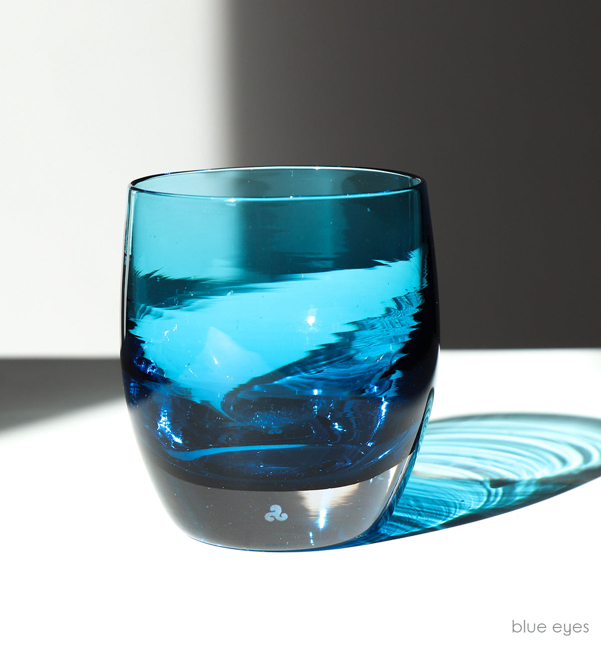 blue eyes transparent crystal blue hand-blown glass votive candle holder on a white surface sitting in sunlight, with its shadow glistening behind.