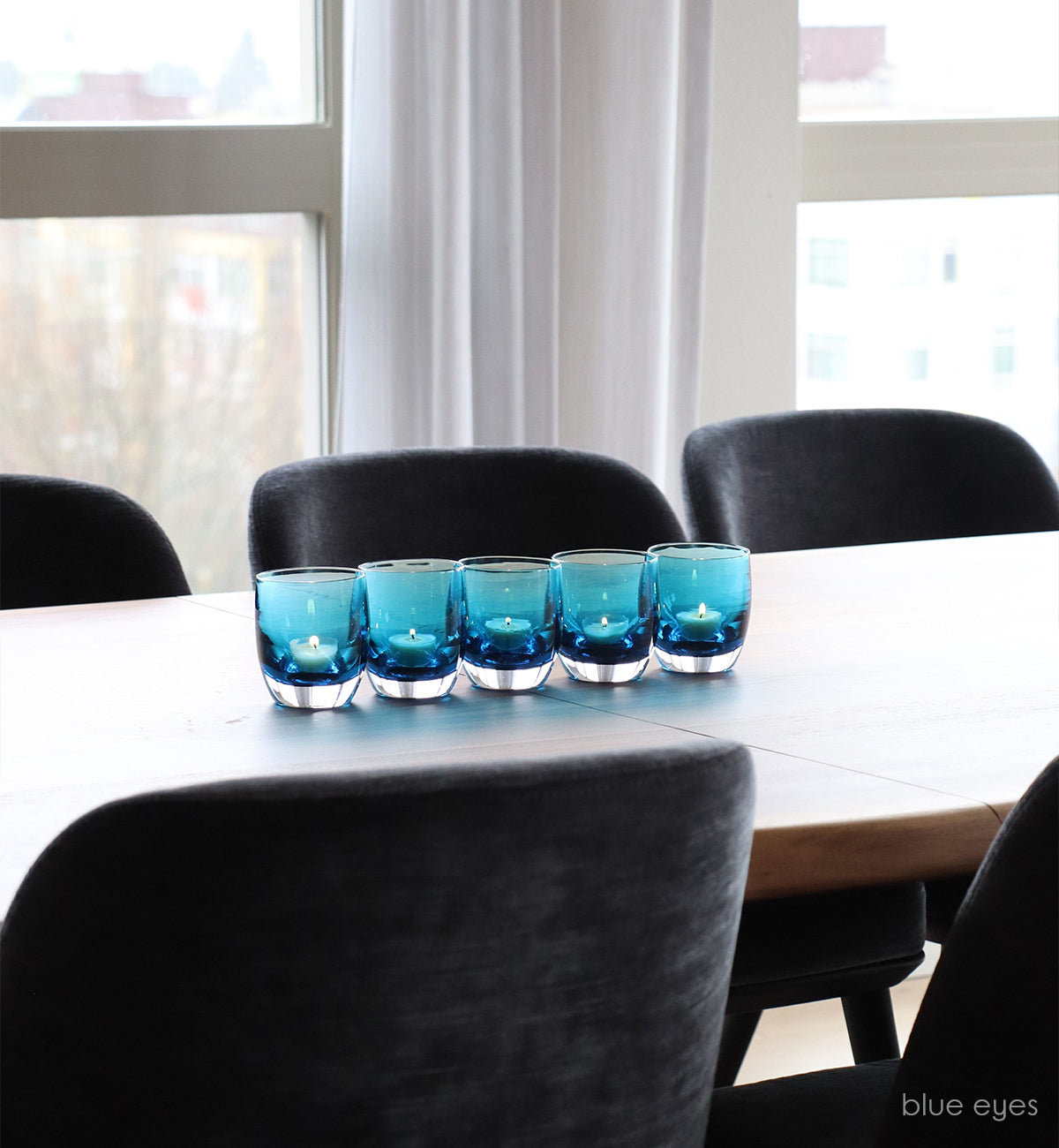 five blue eyes transparent crystal blue hand-blown glass votive candle holders on a wood dining room table with charcoal velvet dining chairs.