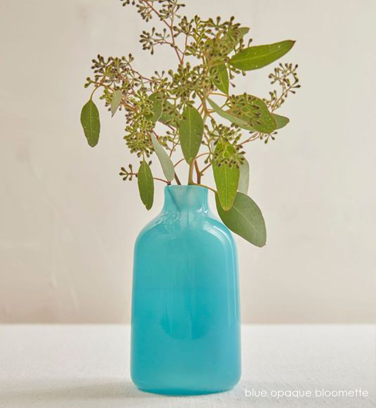 blue opaque bloomette, blue opaque hand-blown glass flower bud vase with green plant inside on a cream table.