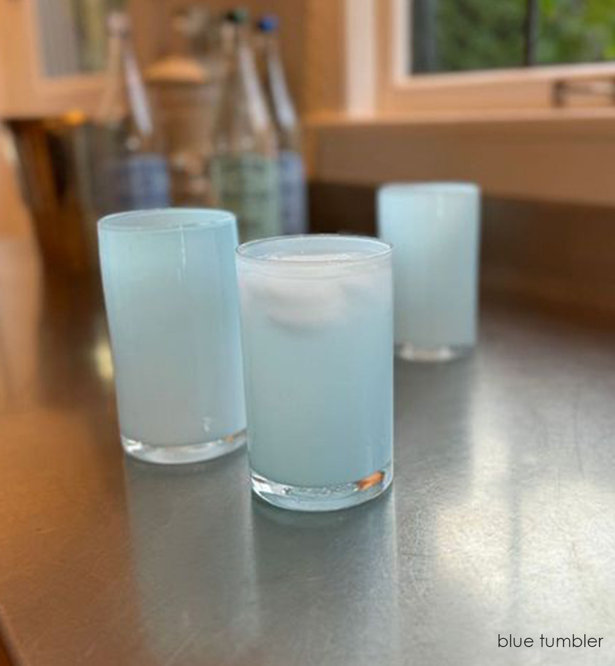 three blue tumblers, light opaque ocean blue hand-blown water glasses with liquid and ice in each on a kitchen counter with sparkling water and a copper ice bucket in background.