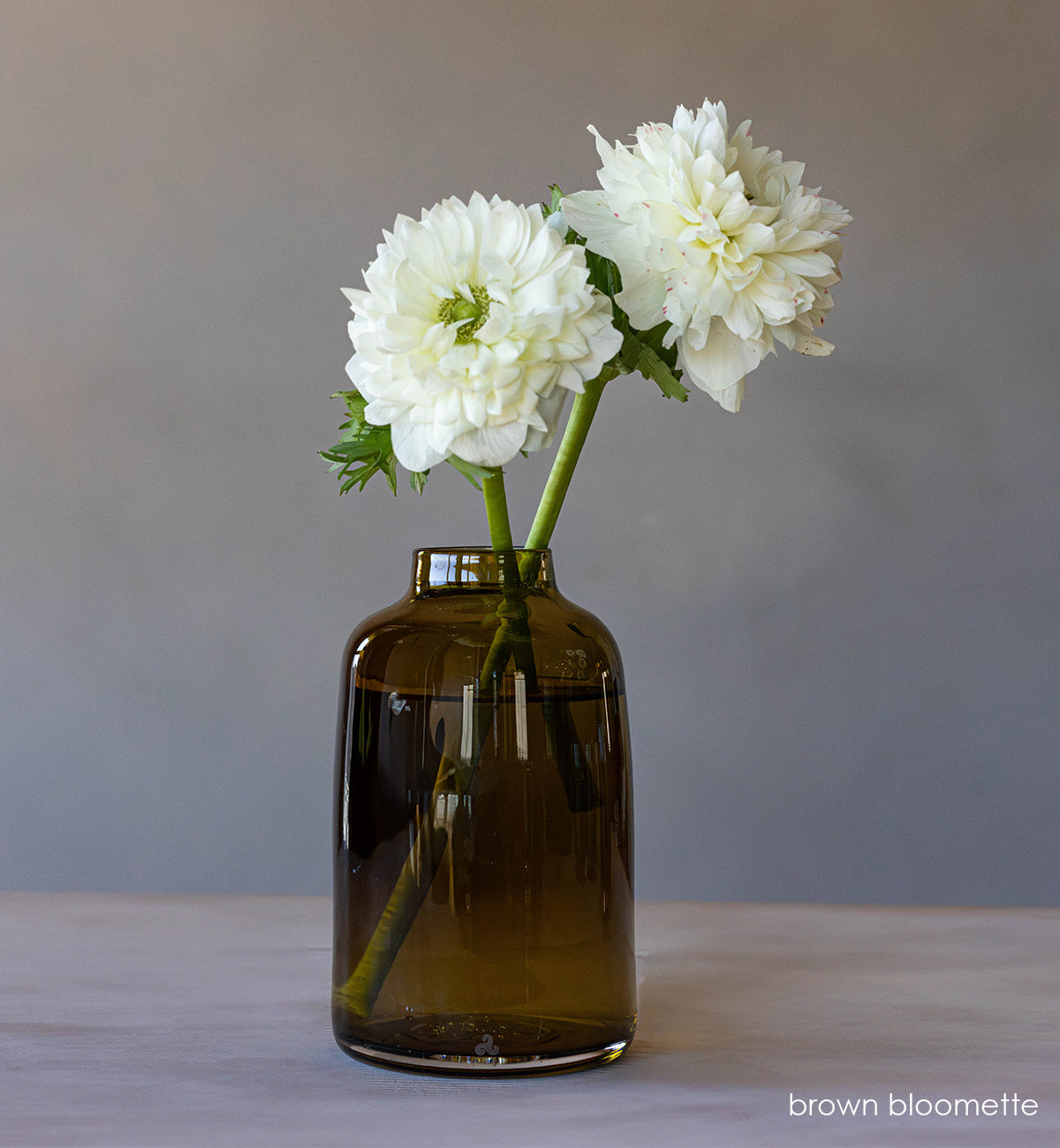 transparent brown hand-blown glass flower bud vase with white flowers on wood table and gray background