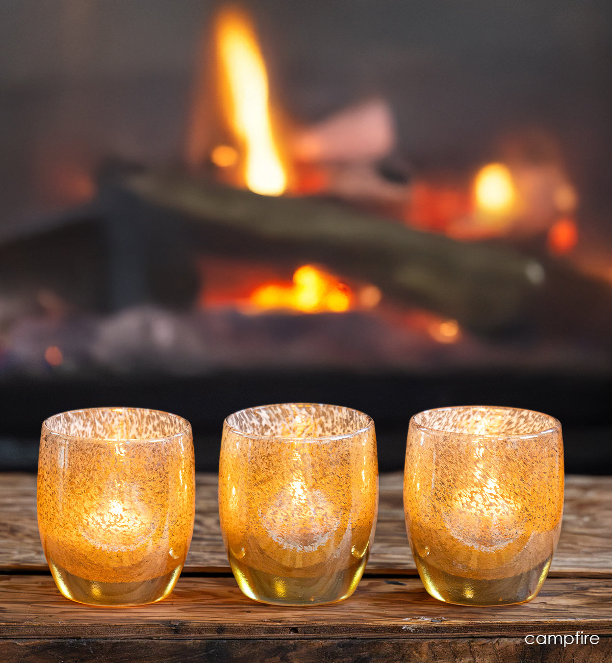 three campfire, gold speckled glassybaby, hand-blown glass candle holders in front of a fire.