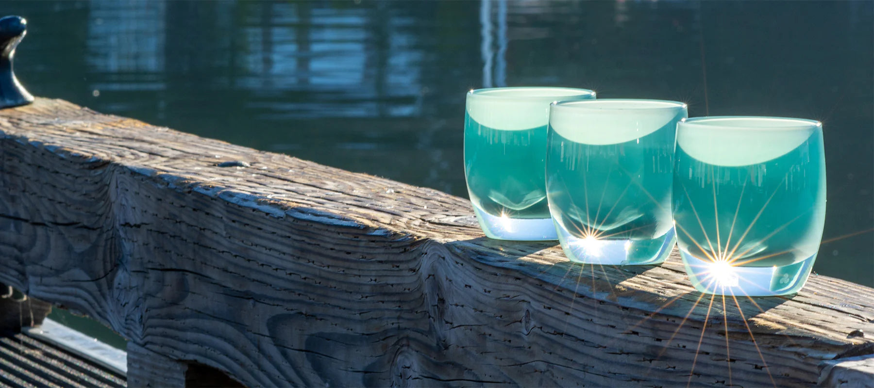 three begin again, light teal, hand-blown glass votive candle holders on a wood perch overlooking water.
