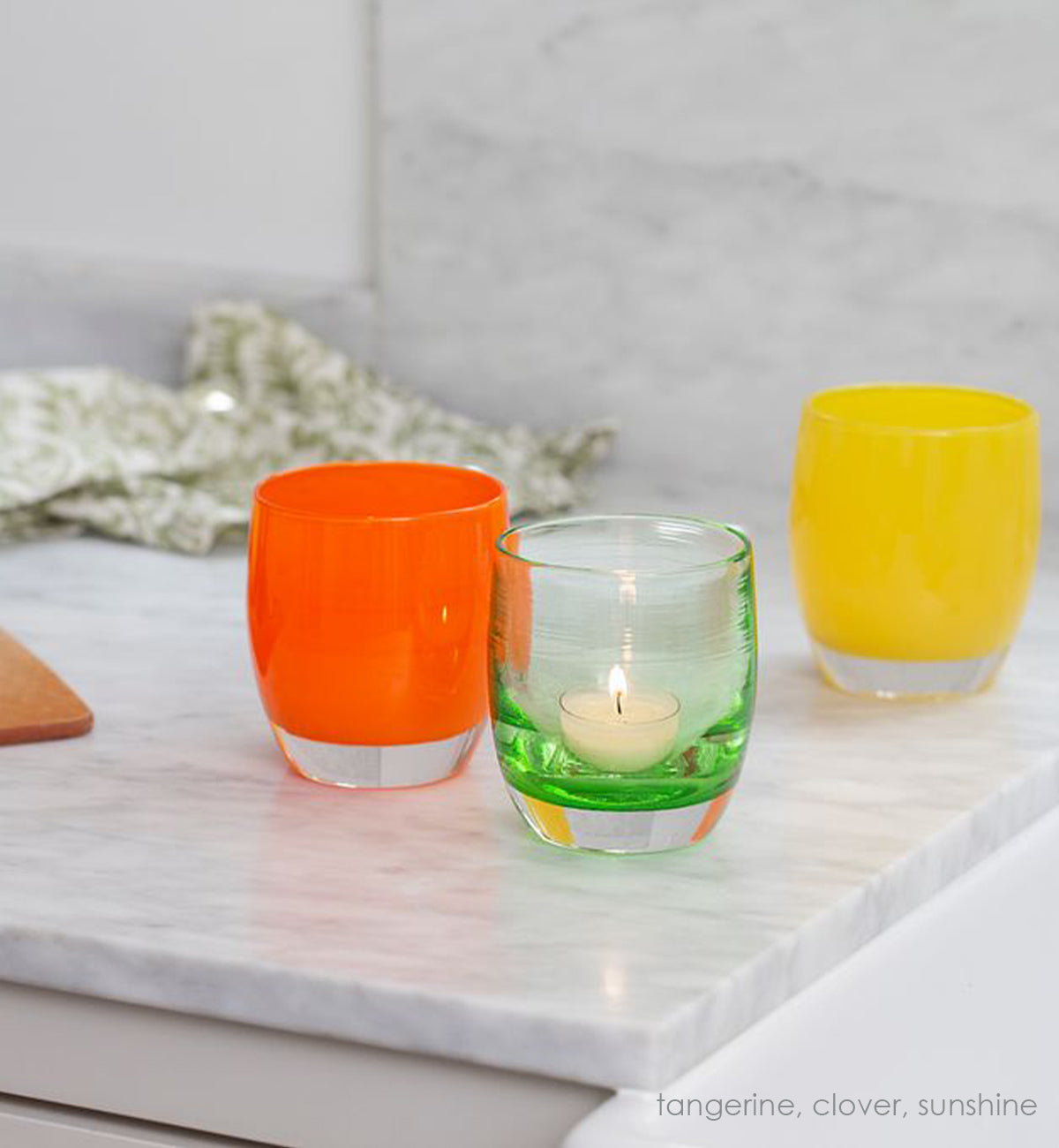 clover, transparent light green hand-blown glass votive candle holder with tangerine and sunshine on a white and gray marble counter top next to a wood cutting board and green patterned dish towel.