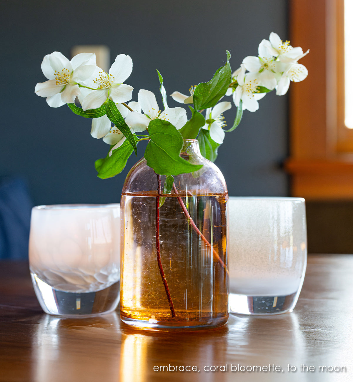 coral bloomette , transparent coral hand-blown glass flower bud vase white flowers and two white embrace and to the moon glassybaby next to it on a wood table