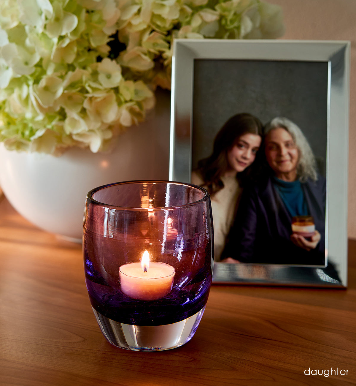 'daughter' lit purple hand-blown glass candle holder on wood dresser. Photo of mother and daughter in silver photo frame and flowers in white vase in background.