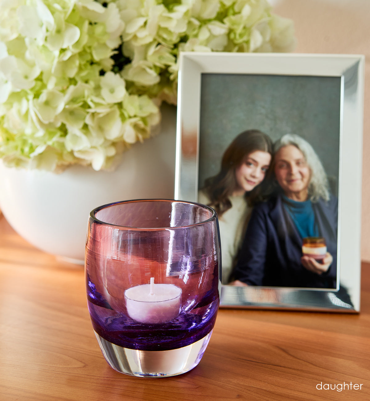'daughter' purple hand-blown glass candle holder on wood dresser. Photo of mother and daughter in silver photo frame and flowers in white vase in background.