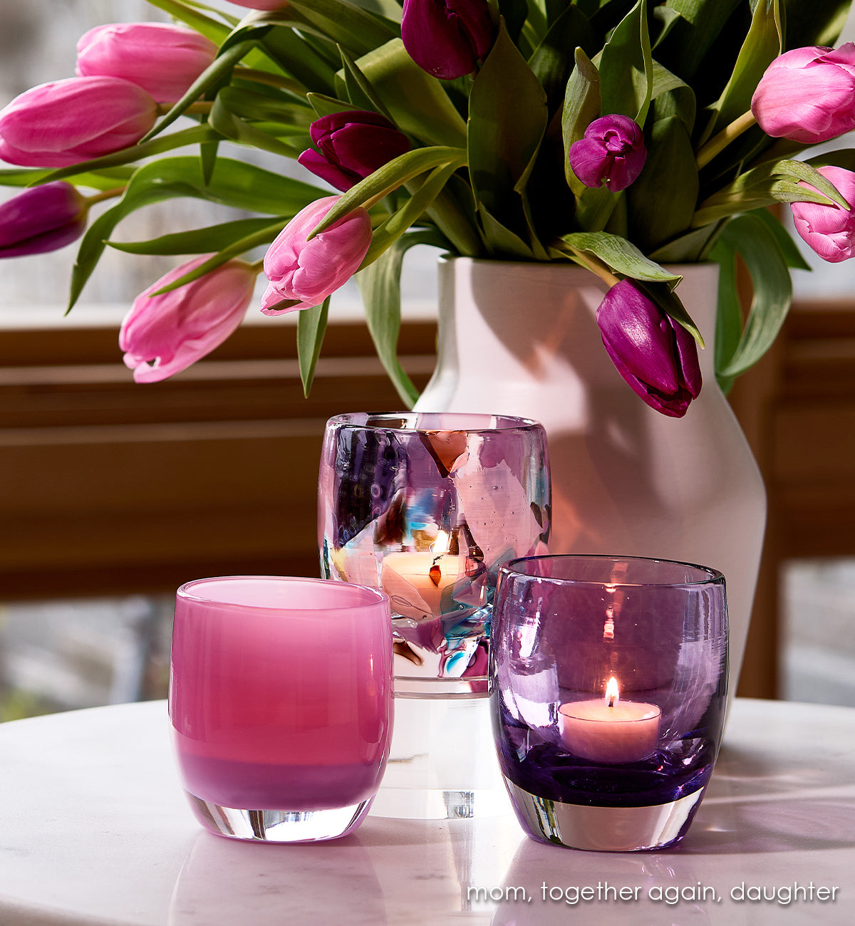 mom, together again, daughter Pink and purple glass candle holders with a vase of pink tulips on a table.