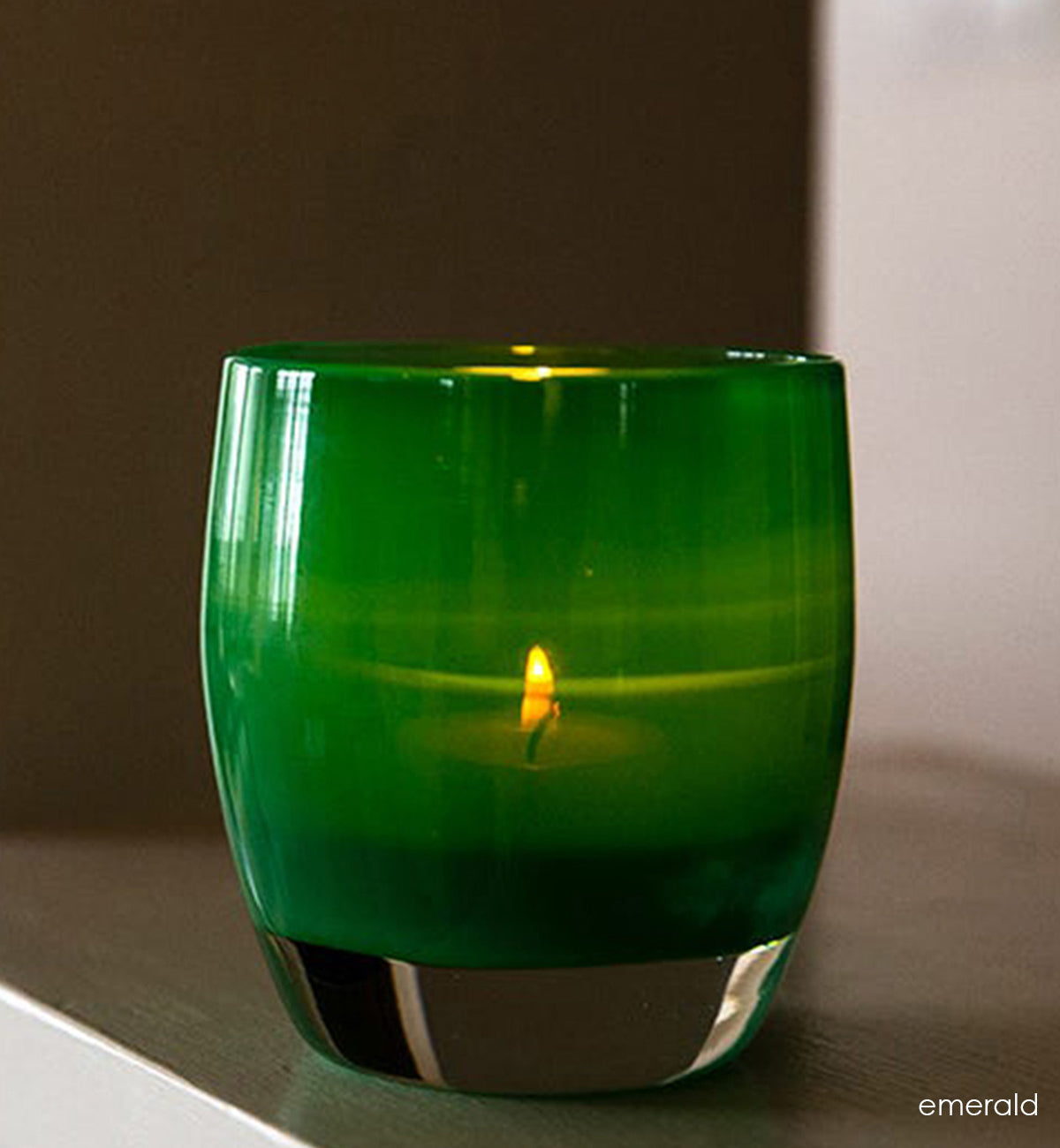 emerald is green a hand-blown glass votive candle holder on light wood shelf.