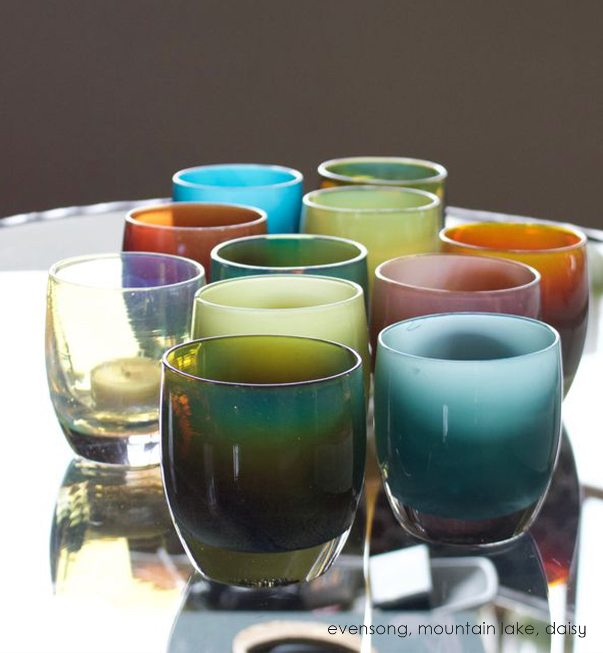 evensong hand-blown green, blue and brown glass votive candle holder in a grouping with mountain lake and daisy on a reflective glass coffee table.