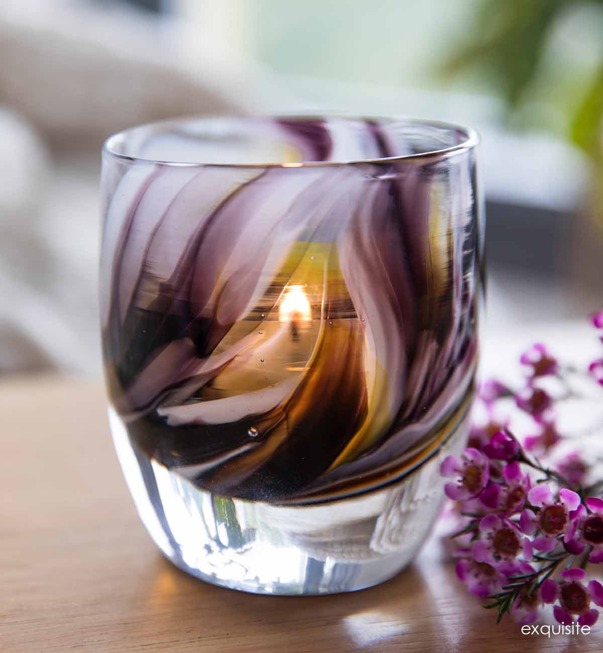exquisite, purple gray yellow swirl, hand-blown glass votive candle holder on a light wood surface with purple flowers.