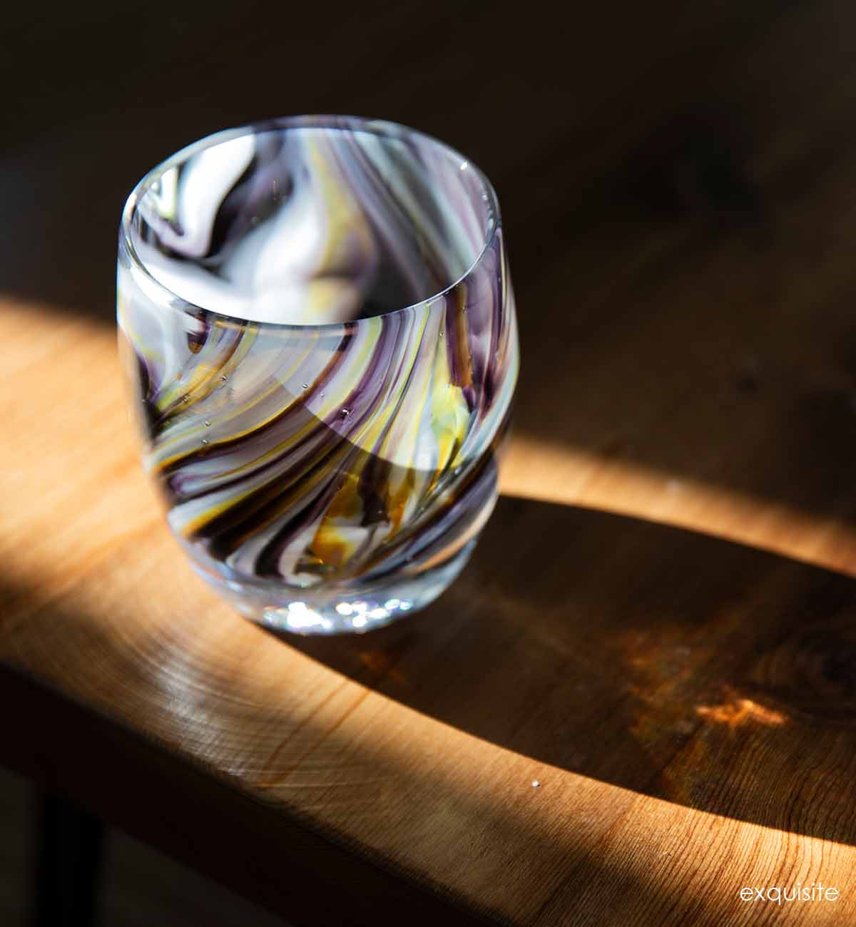 exquisite, purple gray yellow swirl, hand-blown glass votive candle holder sitting in the sunlight on a light wood table.