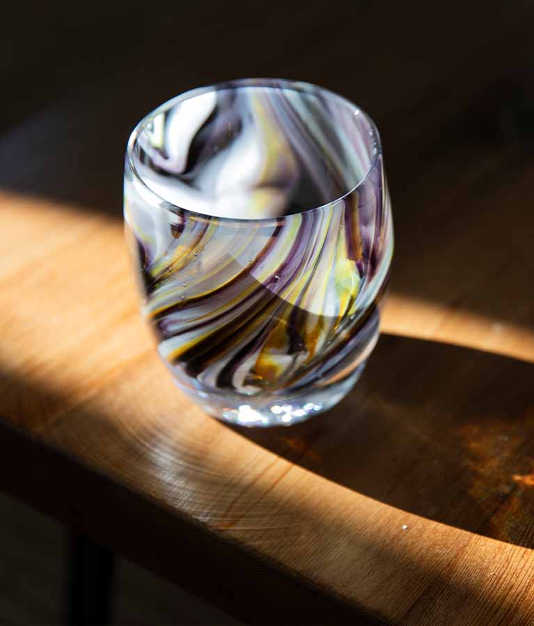 exquisite sitting on a wooden tabletop with sun sining through it, purplish gray swirled with yellow and white, hand-blown glass votive candle holder. 