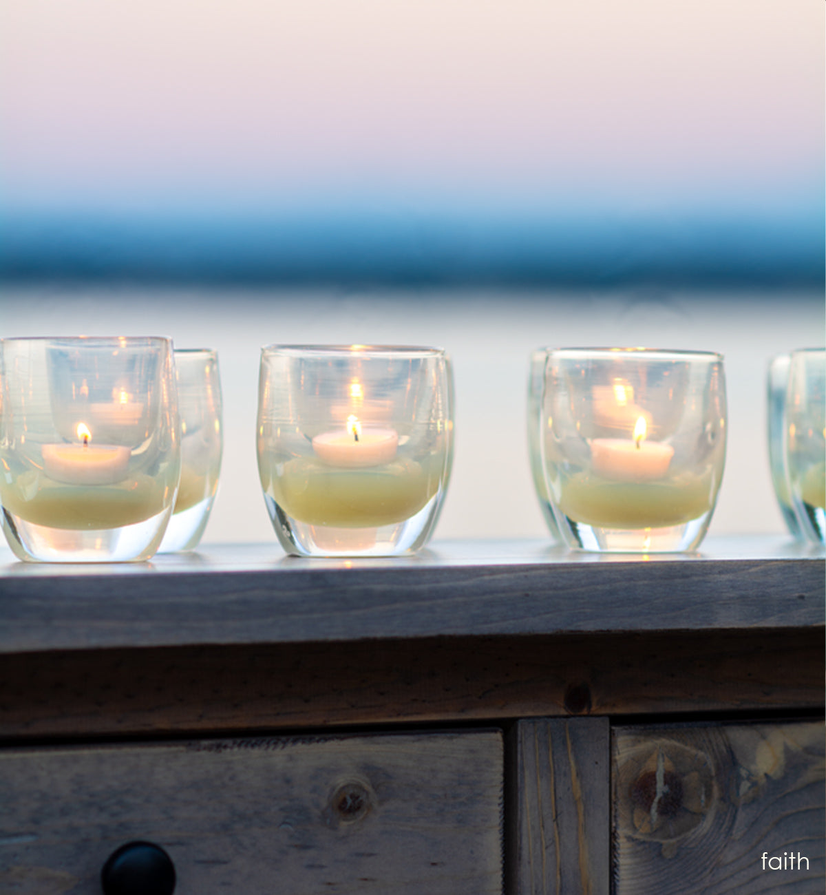 several faith translucent cream hand-blown glass votive candle holders on a wood dresser overlooking ocean water and the sunset.