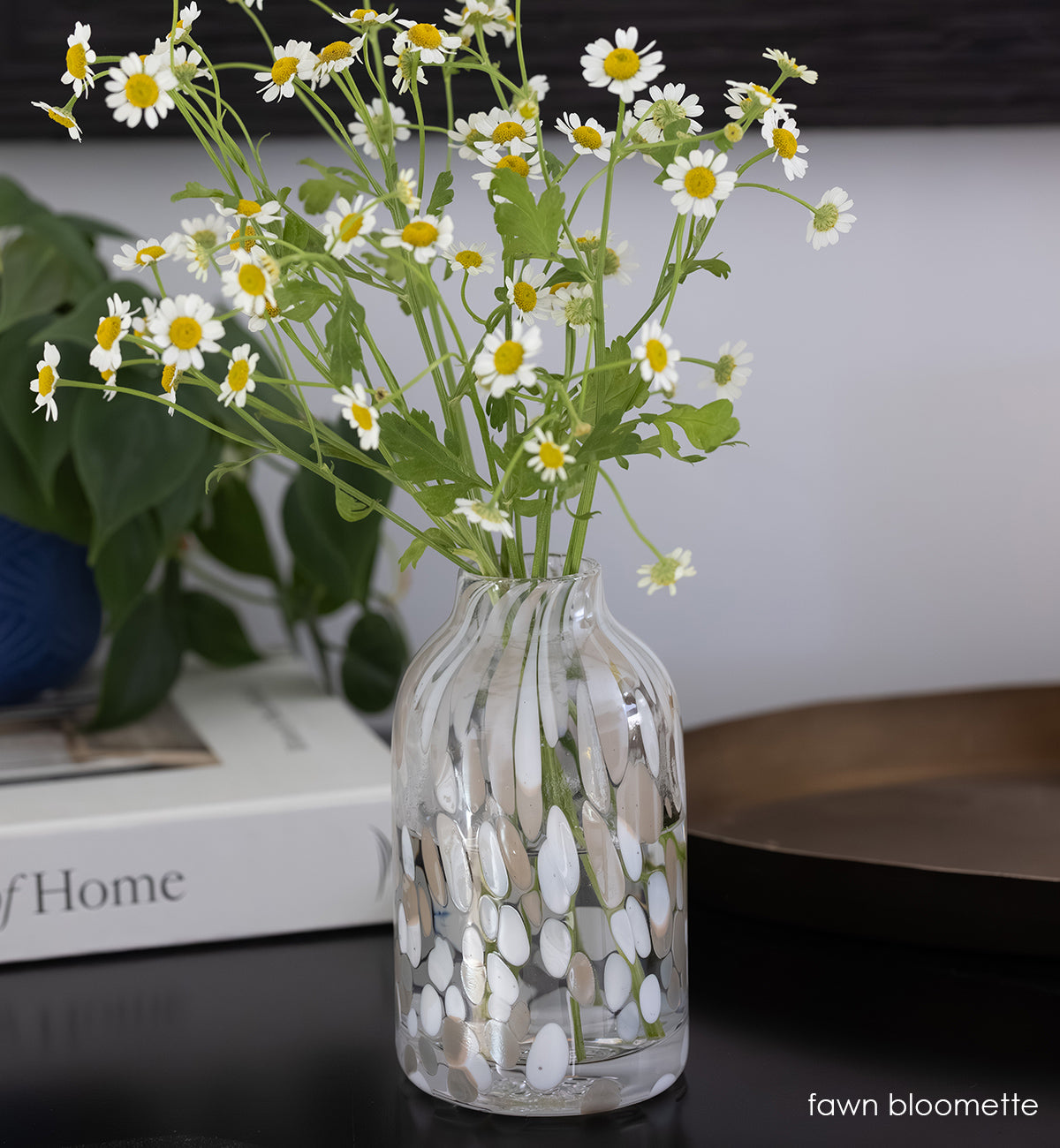 clear bloomette vase with white and tan specs holding a bouquet of white and yellow flowers on a dark surface.
