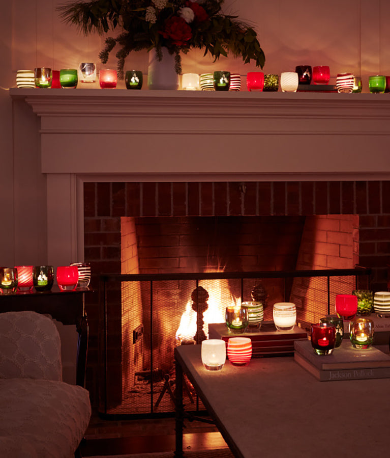 glassybaby holiday collection in living room: on side tables, on coffee table, on mantle with lit fire in fireplace.