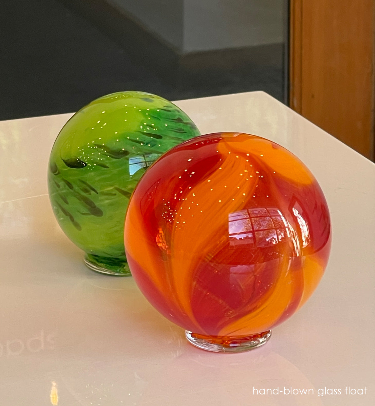 Two hand-blown glass floats, one green and one red-orange, on a reflective surface.