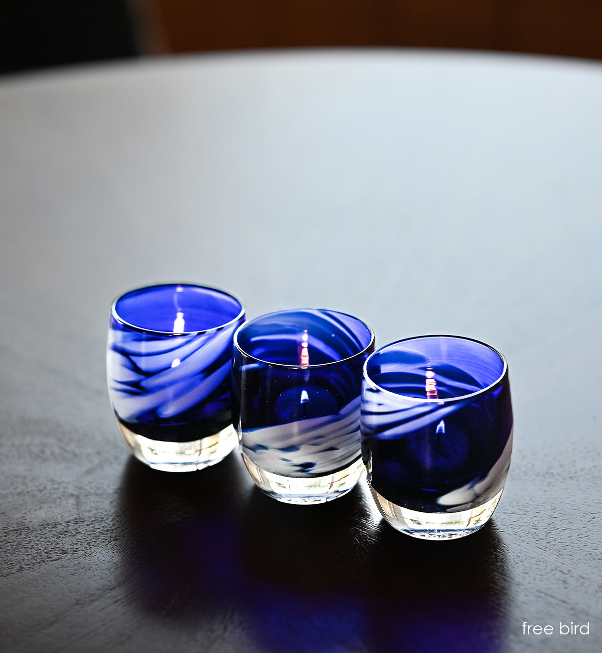 three freebird blue with white wing, hand-blown glass votive candle holders on dark wood circular table.