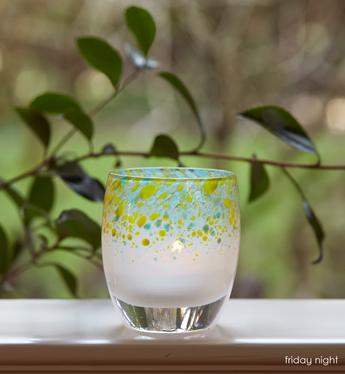 friday night blue green topped hand-blown glass votive candle holder, on a windowsill looking over a green yard.