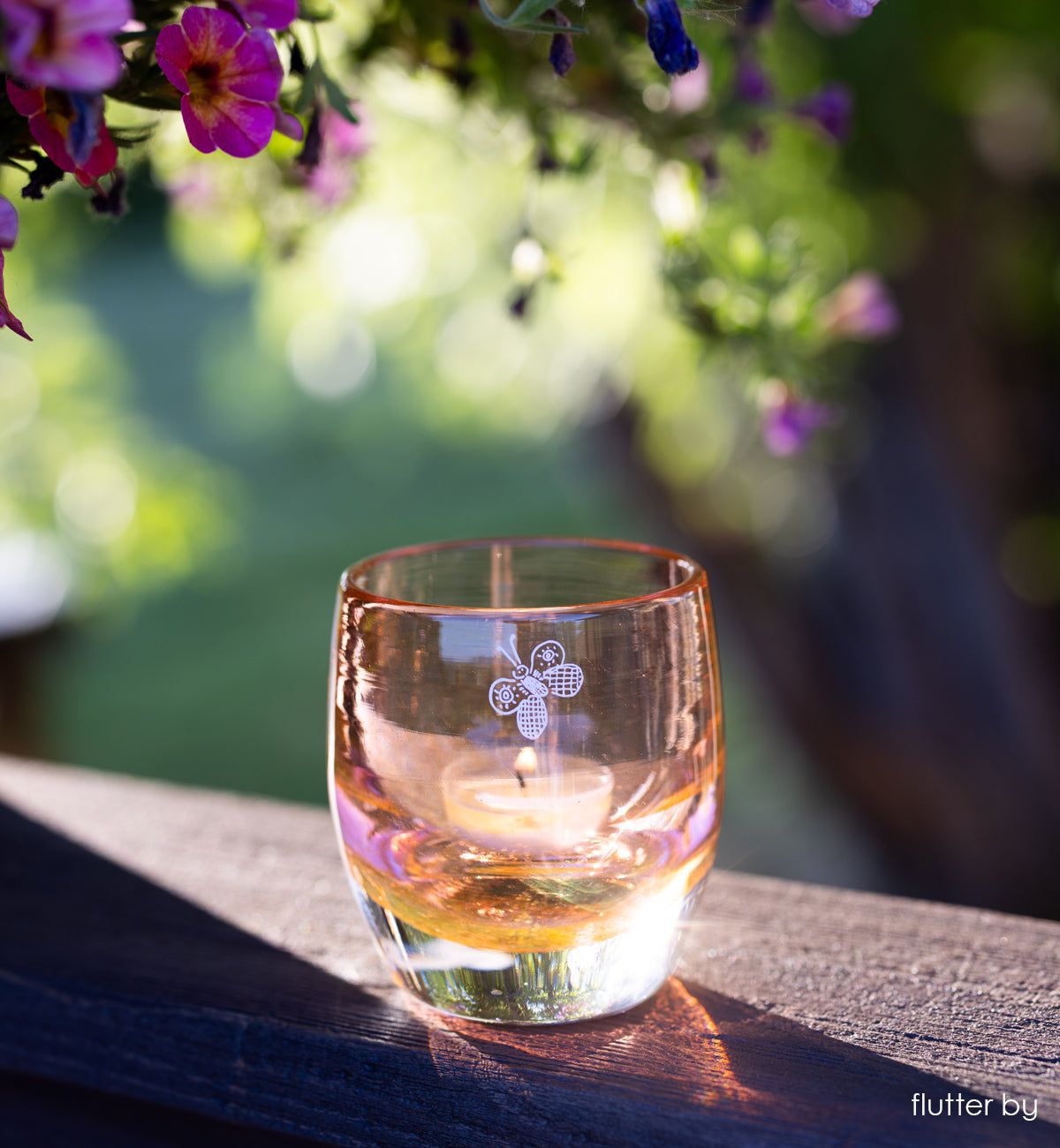 'flutter by' transparent rusty orange with engraved butterfly hand-crafted glass candle holder on wood porch railing with sun shining through. purple flowers in the background.