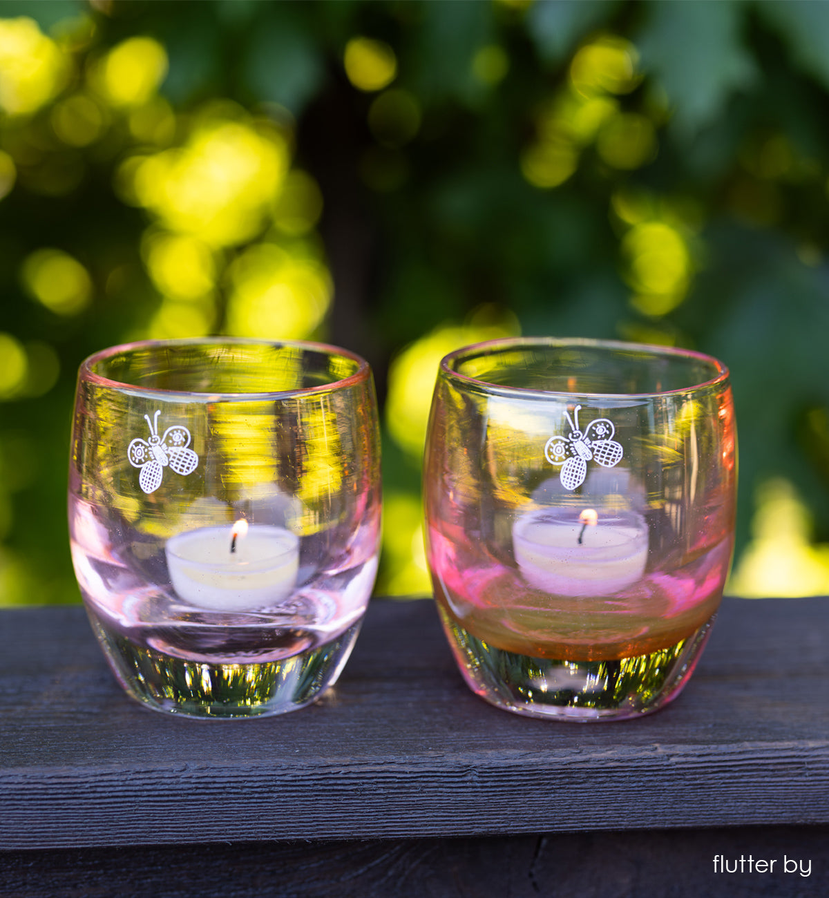 two 'flutter by' transparent rusty orange with engraved butterfly hand-crafted glass candle holders on wood porch railing and green trees in background.