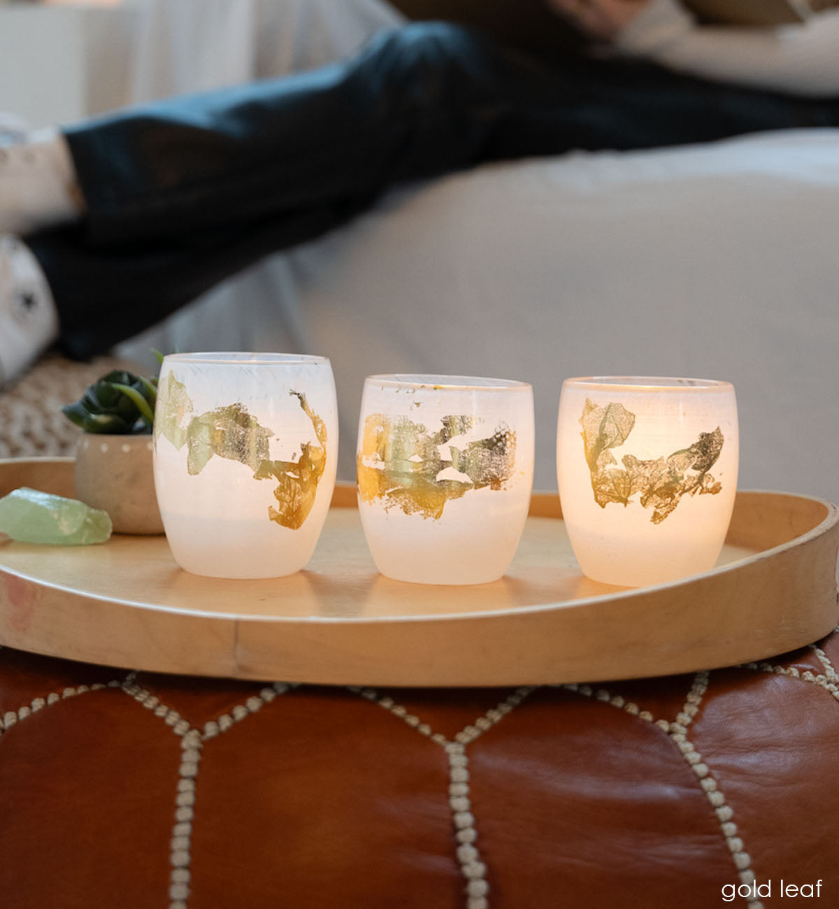three gold leaf white with 24k gold hand-blown glass votive candle holders on a wood tray on a tan ottoman with somebody reading in the background.