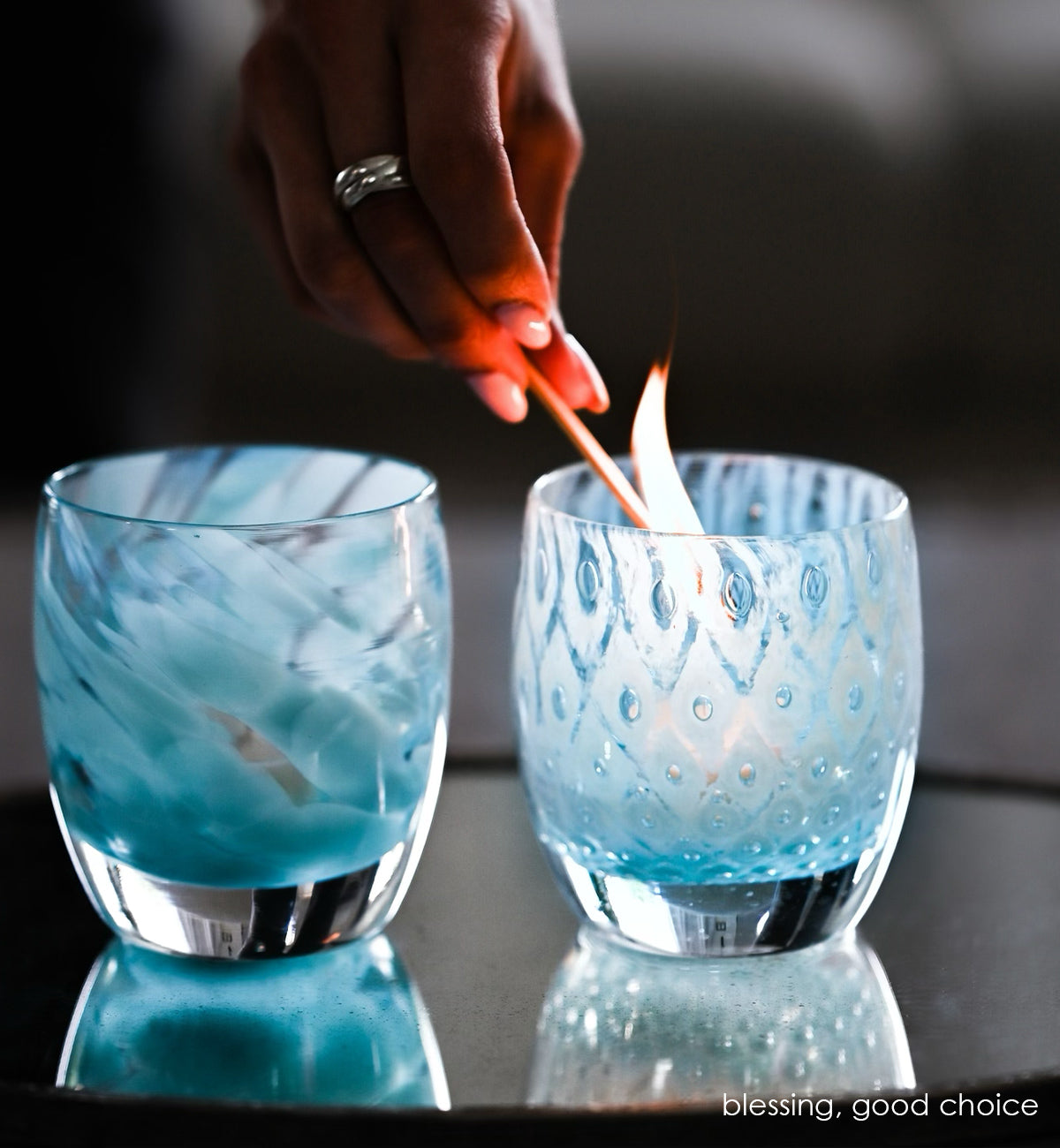 good choice light blue bubble, hand-blown glass votive candle holder being lit with a match next to blessing on a reflective surface.